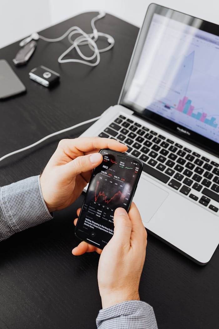 Hands holding smartphone displaying stock charts by a laptop, showing market analysis.